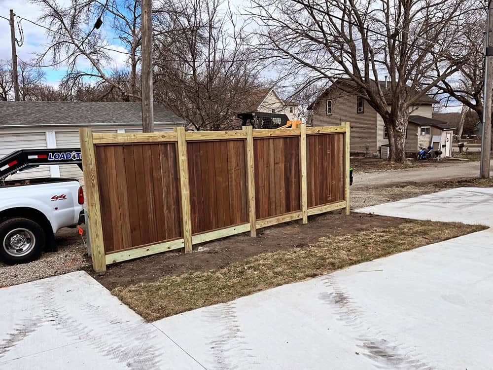 A wooden privacy fence under construction stands on a patch of dirt beside a concrete driveway, with houses, bare trees, and utility poles visible in the background.