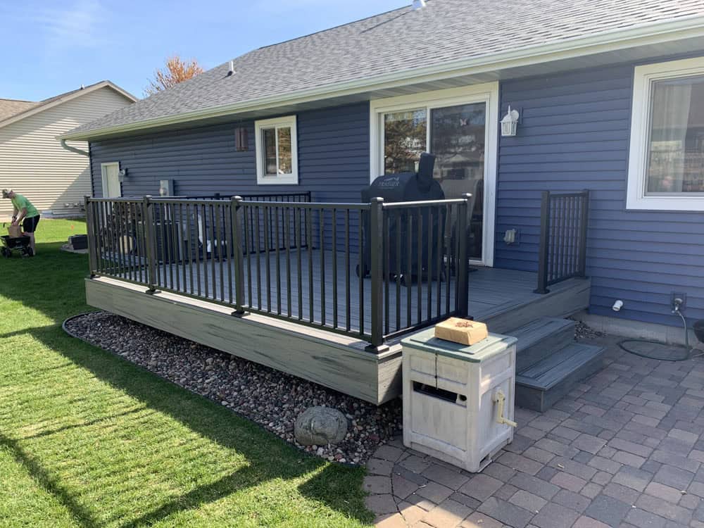 A small backyard deck with gray flooring, black railings, a grill, and a covered trash bin. The deck connects to a blue house and is surrounded by grass and a stone walkway.