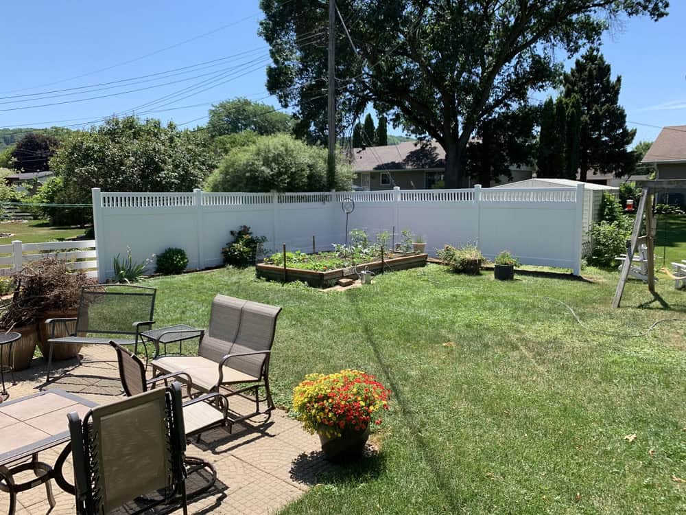 Backyard with a patio set, potted flowers, green lawn, raised garden beds, and a white privacy fence; trees and houses are visible in the background under a clear blue sky.
