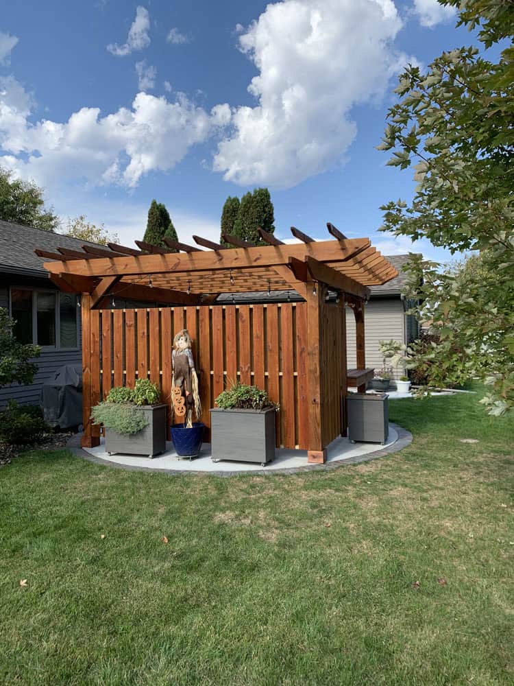 A wooden pergola stands on a circular concrete pad in a grassy backyard, surrounded by potted plants. A carved wooden statue is displayed inside, with a house and trees in the background under a partly cloudy sky.