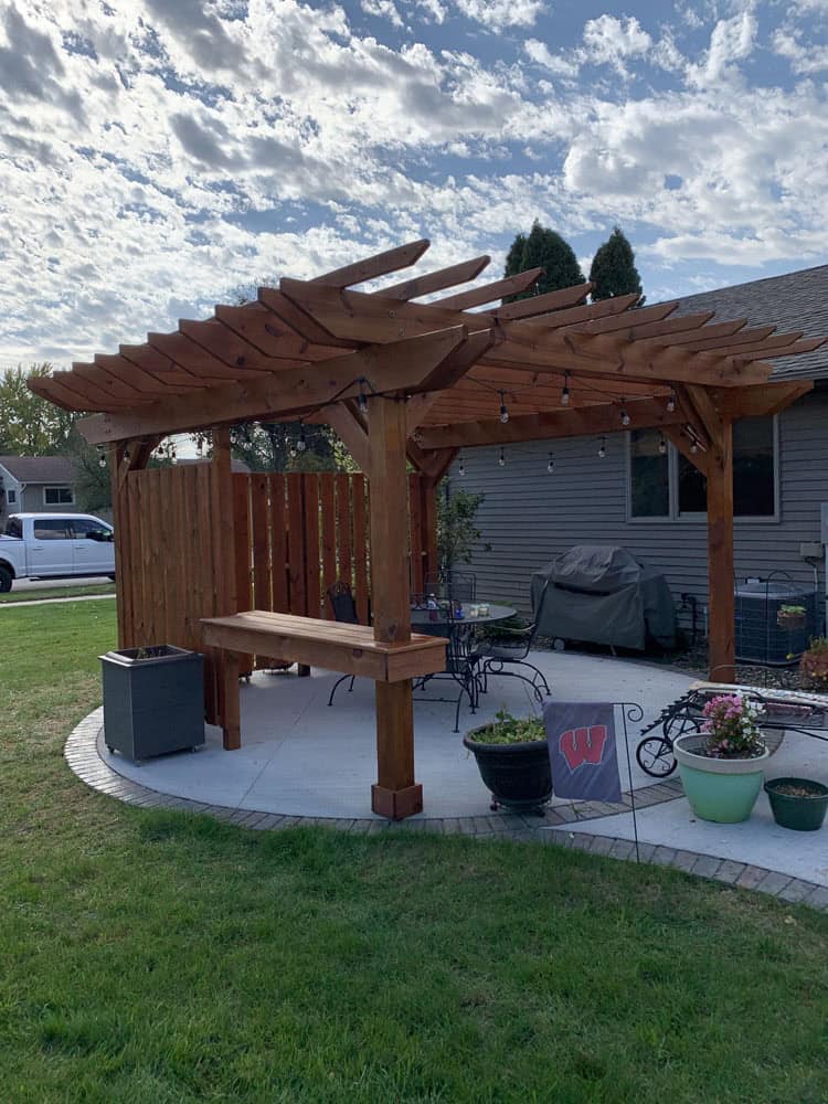 A wooden pergola stands on a tiled patio next to a house, with string lights, potted plants, patio furniture, and a grill underneath. The sky is partly cloudy, and the lawn surrounds the patio.