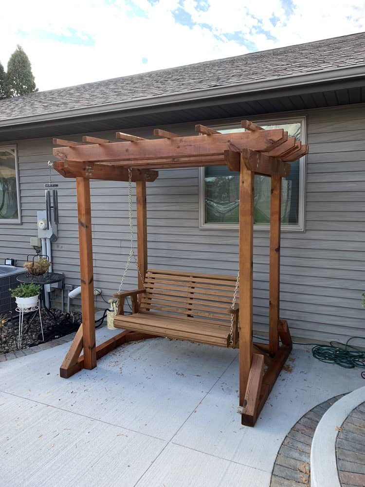 A wooden porch swing with a pergola-style canopy sits on a concrete patio next to a house with gray siding. The swing is suspended by chains and the patio is bordered by garden beds and a garden hose.