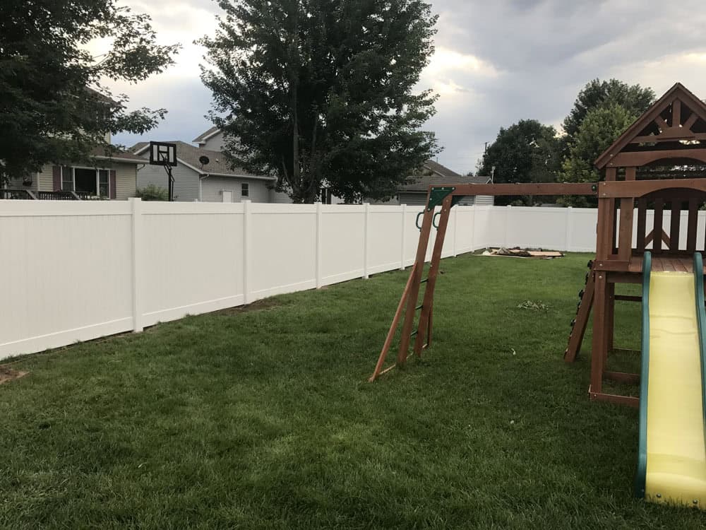 A backyard with green grass, a wooden playset with a yellow slide, and a tall white vinyl privacy fence. Trees and houses are visible in the background under a cloudy sky.