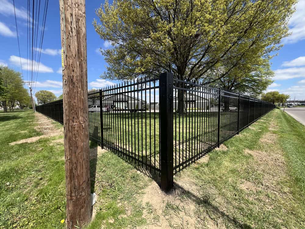 A black metal fence forms a sharp corner on a grassy area with a tree inside, next to a wooden utility pole and a road under a blue sky with scattered clouds.