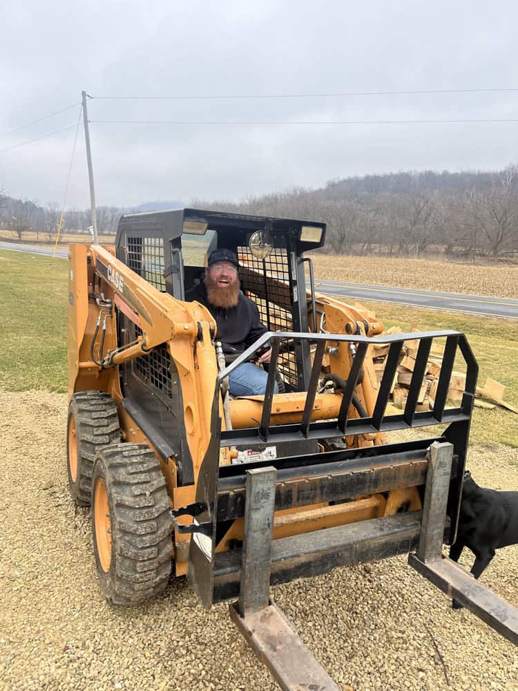 A smiling man with a beard and a cap sits in the driver’s seat of an orange Case skid steer loader on a gravel surface, with grassy fields and trees in the background.