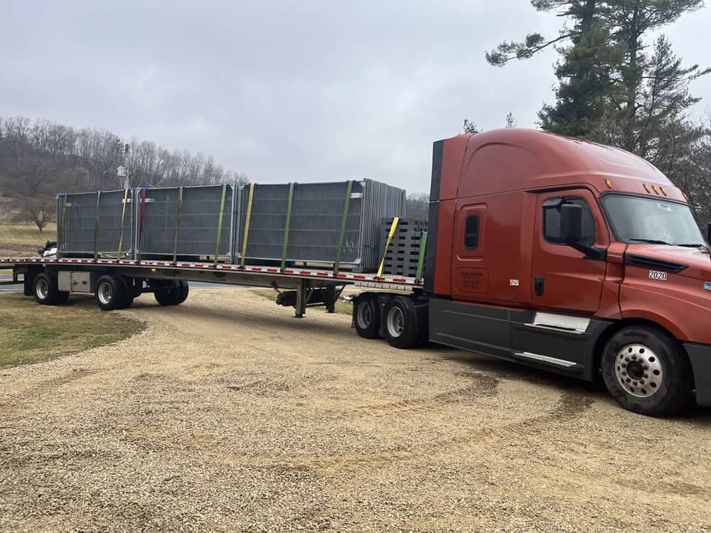 A red semi-truck with a flatbed trailer is parked on a gravel road, carrying large metal crates secured with straps. Trees and a grassy field are visible in the background under a cloudy sky.