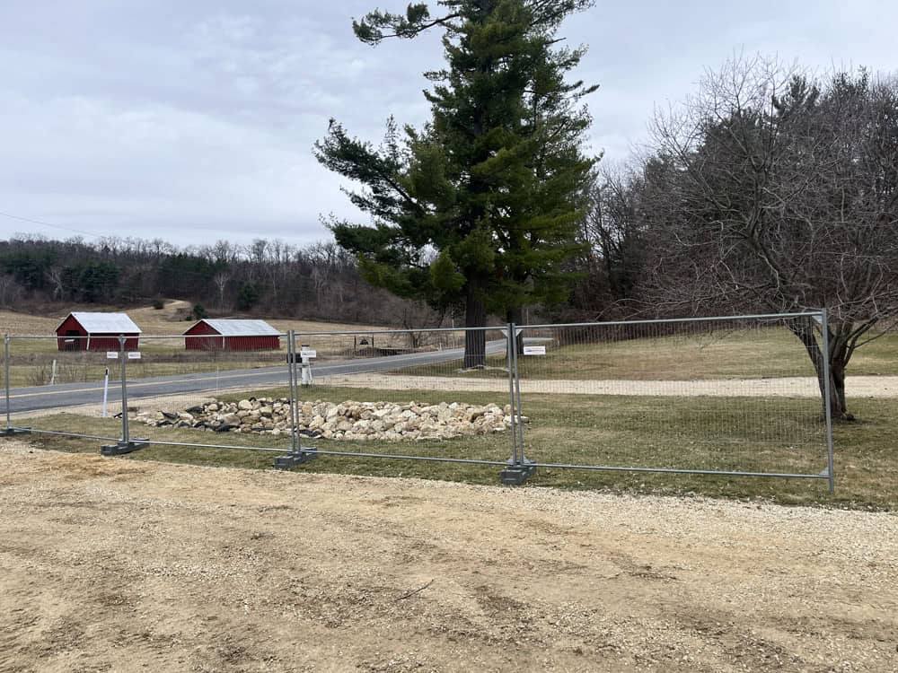 A fenced-off area with rocks and grass is in the foreground, with a gravel path and a large pine tree nearby. Two red barns sit across the road in a rural landscape under a cloudy sky.