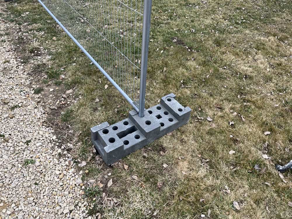 A temporary metal fence is secured at the base with two gray concrete blocks featuring holes, set on grass near a gravel path.