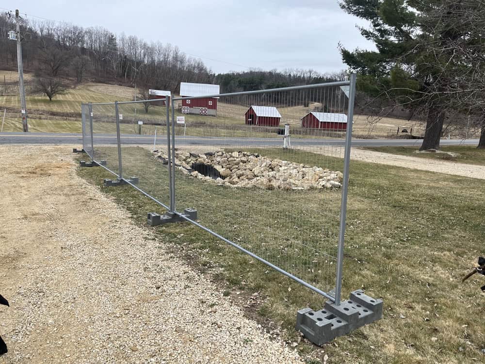 Temporary metal fencing panels set up on a grassy area, with concrete bases supporting each section. In the background, there are red barns, a gravel road, trees, and a rocky drainage area.