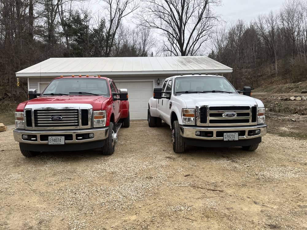 Two Ford pickup trucks, one red and one white, are parked side by side on a gravel driveway in front of a white garage with a metal roof. Leafless trees and a cloudy sky are in the background.