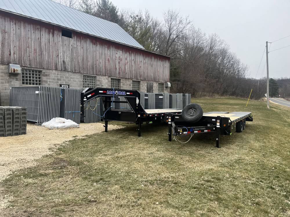 Two flatbed trailers are parked on grass beside a barn with a metal roof. Stacks of fencing materials and wire are visible near the barn. In the background, there are trees and a road runs alongside the property.