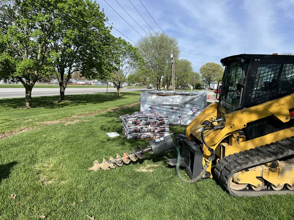A yellow skid steer with an auger attachment drills into grass near a stack of concrete bags and paving stones. Trees and a road are in the background on a sunny day.