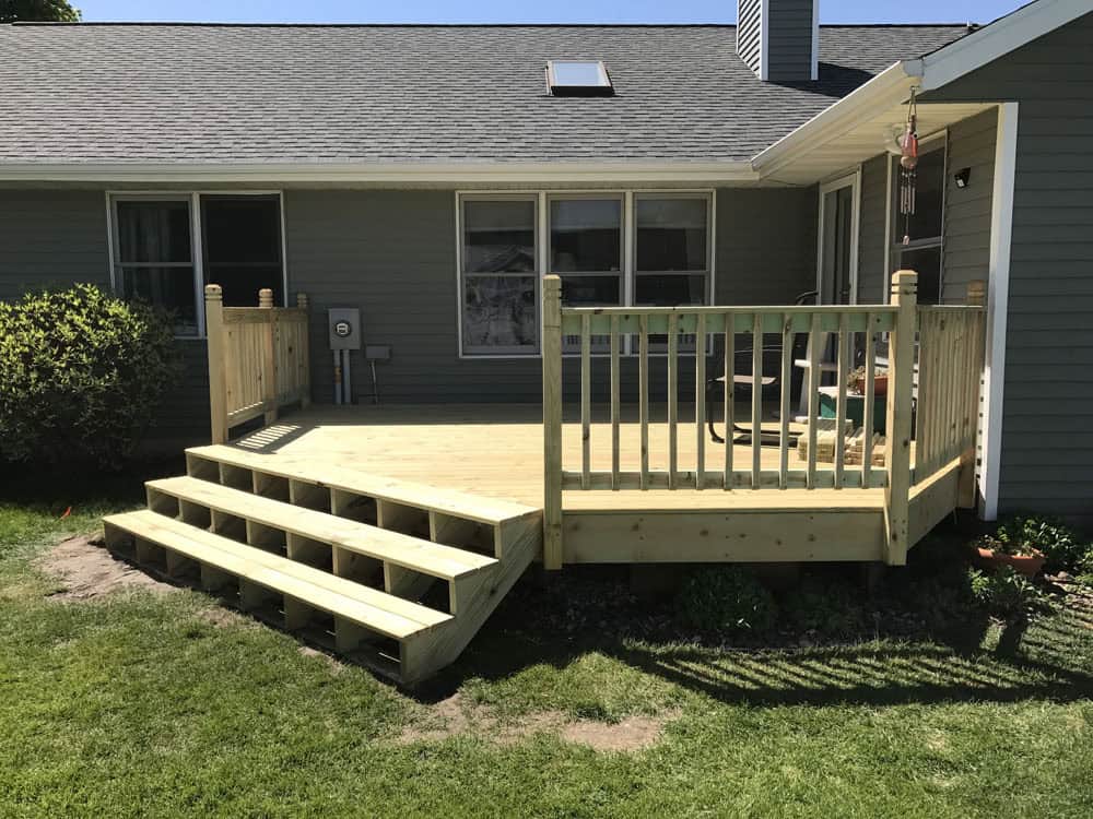 A newly built wooden deck with railings and four steps leading down to a grassy backyard, attached to a gray house with several windows and a small table and chairs on the deck.