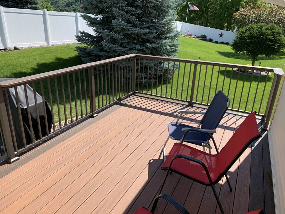 A backyard deck with two chairs, one red and one blue, overlooks a green lawn with trees and a white fence. An American flag is visible in the background.