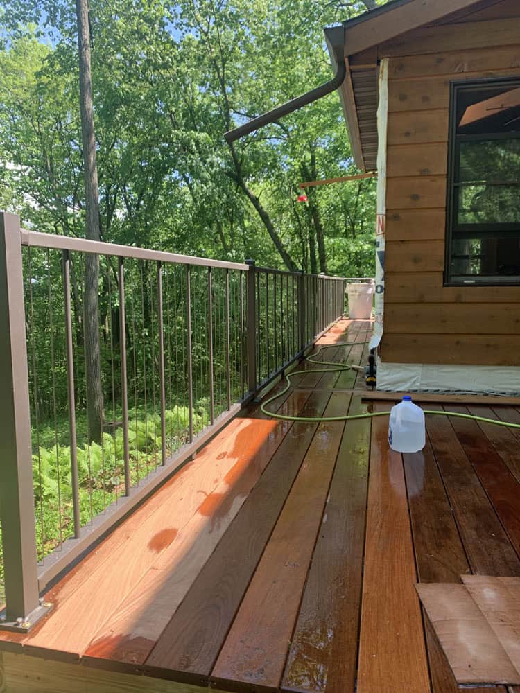 A sunlit wooden deck beside a house with metal railings, surrounded by green trees. A garden hose and a plastic water jug sit on the deck, which appears freshly cleaned or wet in some areas.