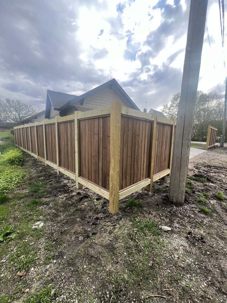 A wooden privacy fence with light-colored posts surrounds a house on a cloudy day. The ground nearby is muddy with patches of green grass, and a utility pole stands in the foreground.