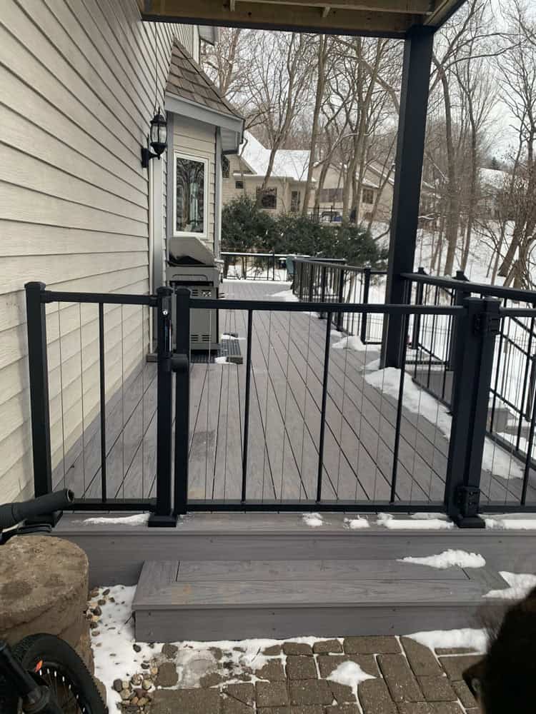 A modern deck with dark metal railings and light gray flooring is attached to a house. Snow patches are visible on the deck, steps, and ground. Leafless trees and houses are in the snowy background.
