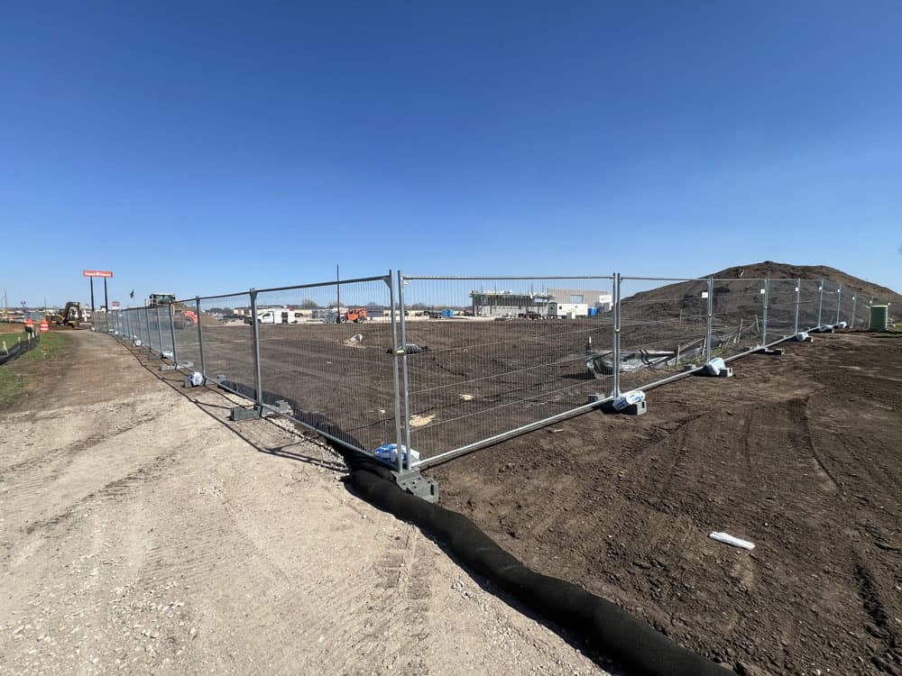 A construction site surrounded by temporary fencing, with dirt piles and some machinery in the background under a clear blue sky. The ground is mostly bare soil, and a few buildings are visible in the distance.