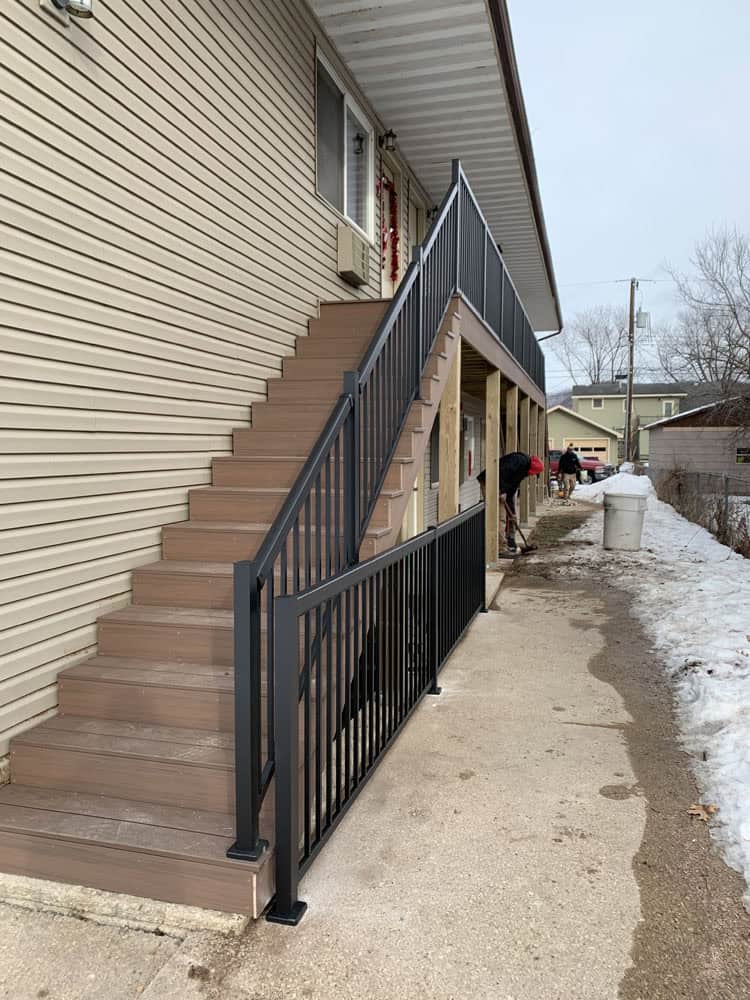 A brown and black exterior staircase with metal railings leads to the second floor of a beige building. Snow is on the ground nearby, and two people are seen shoveling the sidewalk.