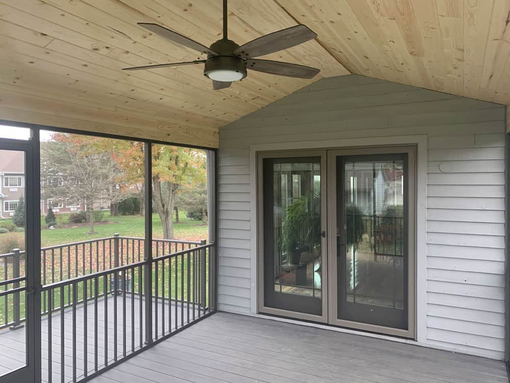 A screened-in porch with a wooden ceiling, ceiling fan, gray siding, double glass doors, and black railing, overlooking a grassy yard with trees and houses in the background.