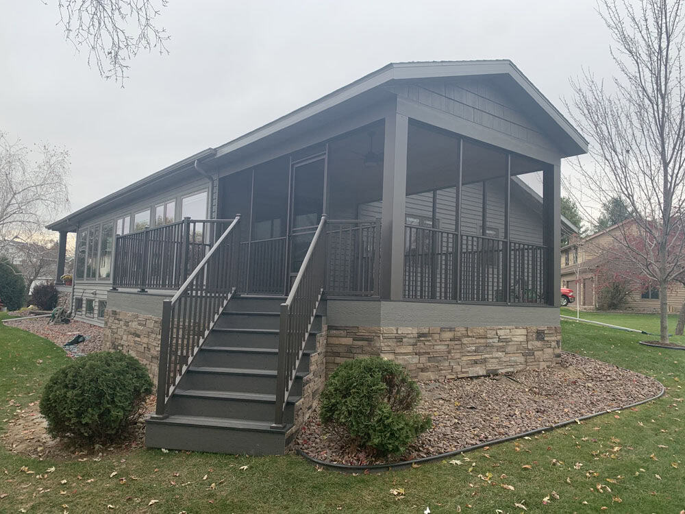 A gray house with a screened-in porch, stone foundation, and wide stairs leading up to the entrance, surrounded by neatly trimmed bushes and a grassy lawn.