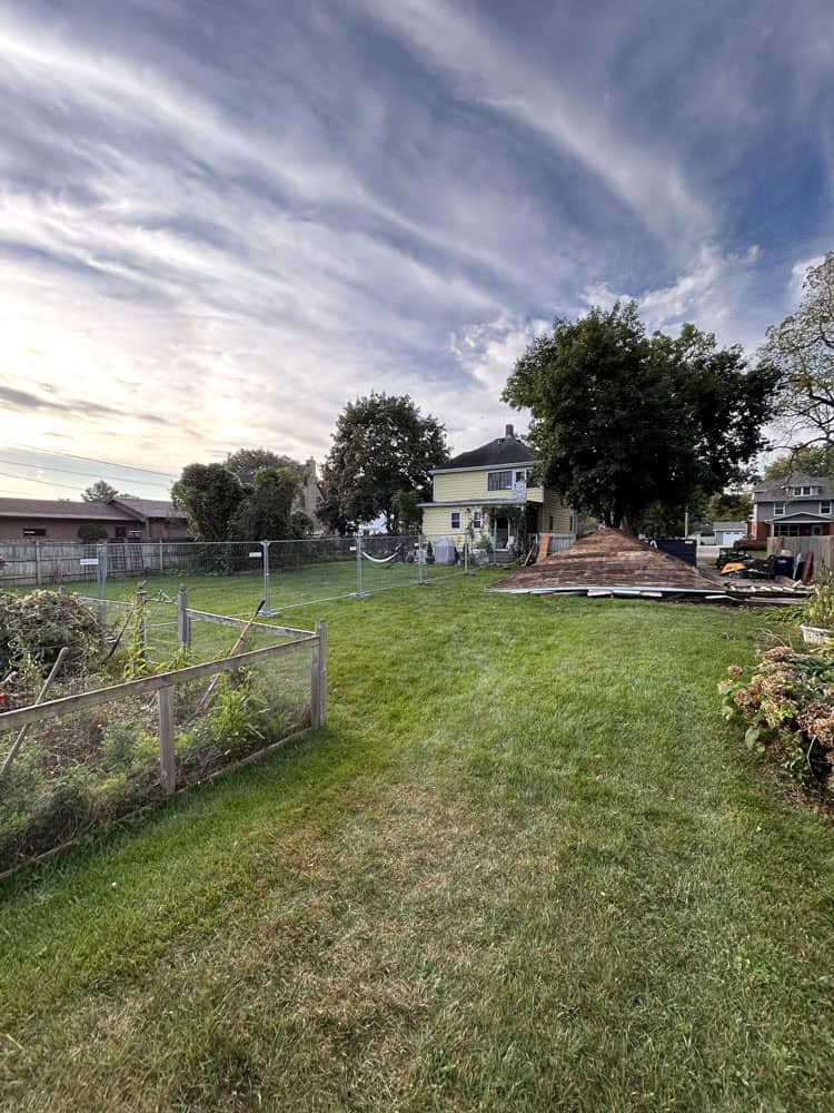 A grassy backyard with a fenced garden area and a house in the background. Construction materials and equipment are on the lawn, and the sky is partly cloudy with streaks of sunlight.