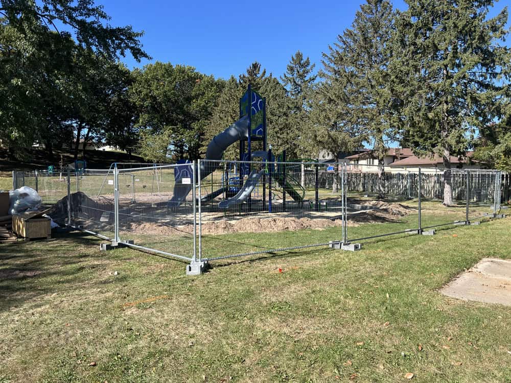 A playground under construction is surrounded by a metal fence. There are slides and climbing structures inside, and patches of dirt. Trees, grass, and houses are visible in the background under a clear blue sky.