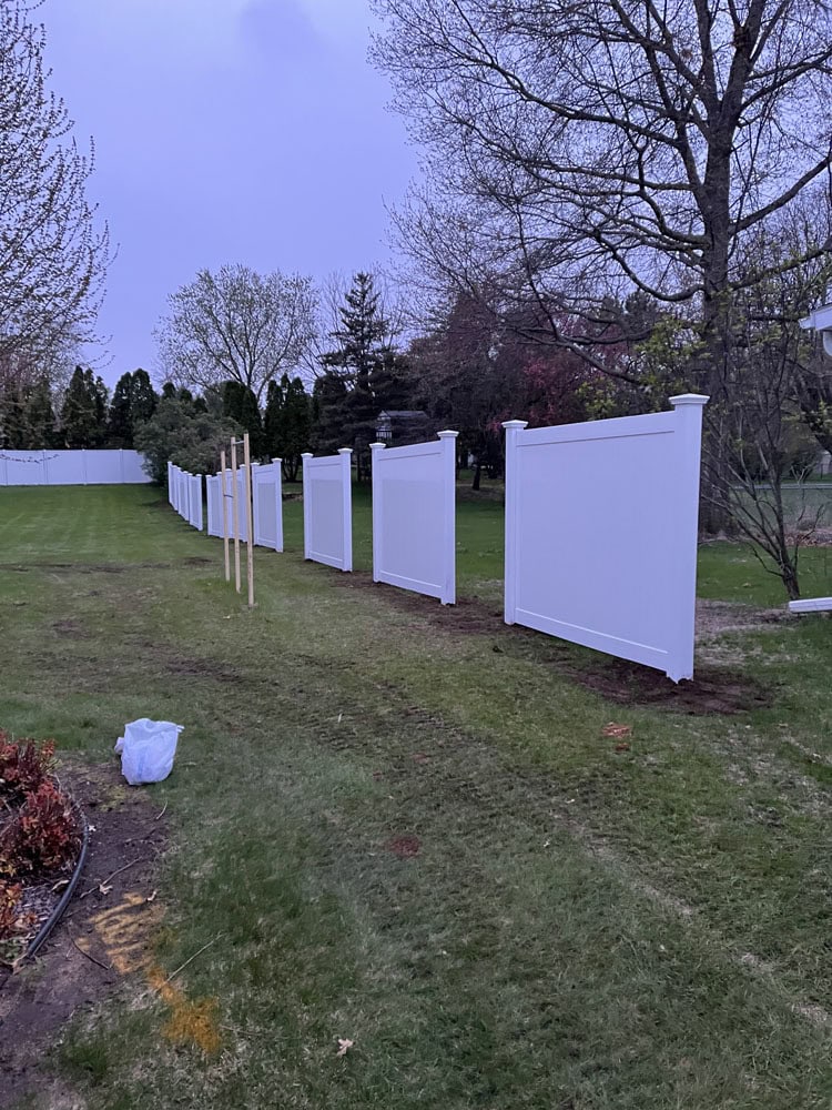 A row of white vinyl fence panels partially installed along a grassy backyard, with wooden stakes marking the alignment and trees visible in the background at dusk.