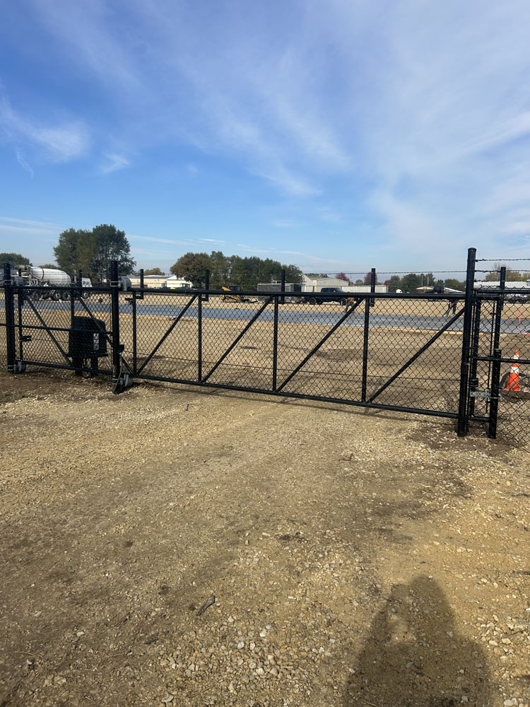 A black chain-link fence gate blocks entry to a gravel area with some orange cones. Beyond the gate is an open, empty lot with trees and industrial buildings in the distance under a blue sky.