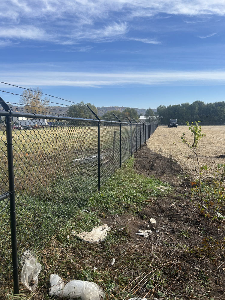 A chain-link fence with barbed wire separates a grassy area from a cleared, dirt-covered field. A vehicle is visible in the distance under a blue sky with wispy clouds. Trash and debris are in the foreground.