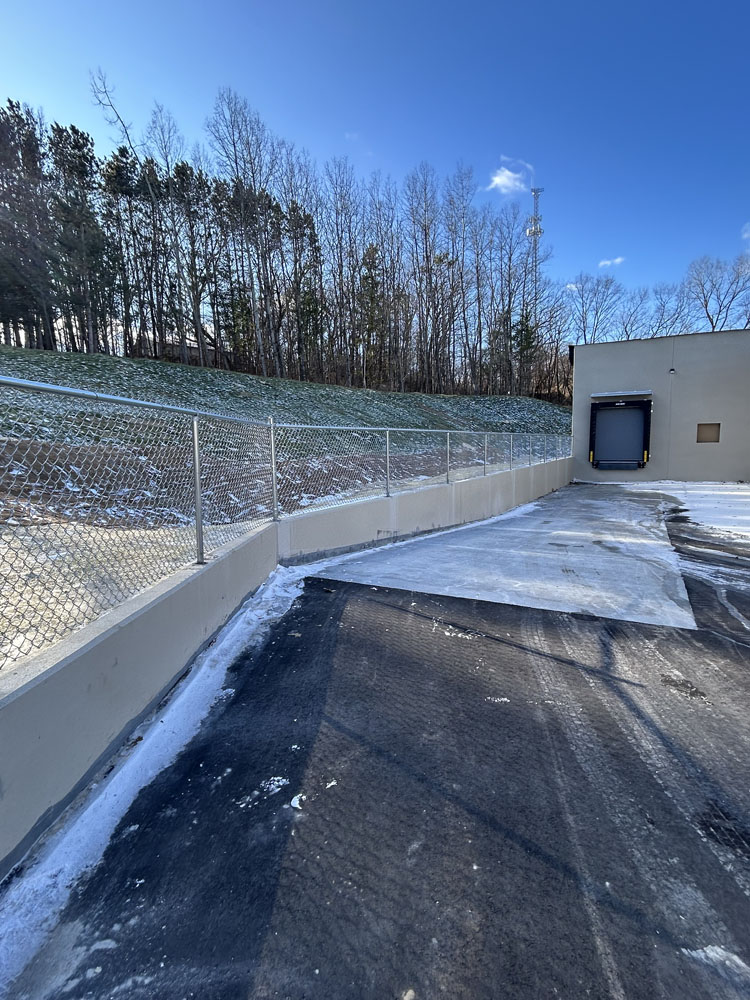A loading dock with a closed bay door sits at the end of a paved area bordered by a chain-link fence and sloped, snow-dusted ground, with leafless trees in the background under a clear blue sky.
